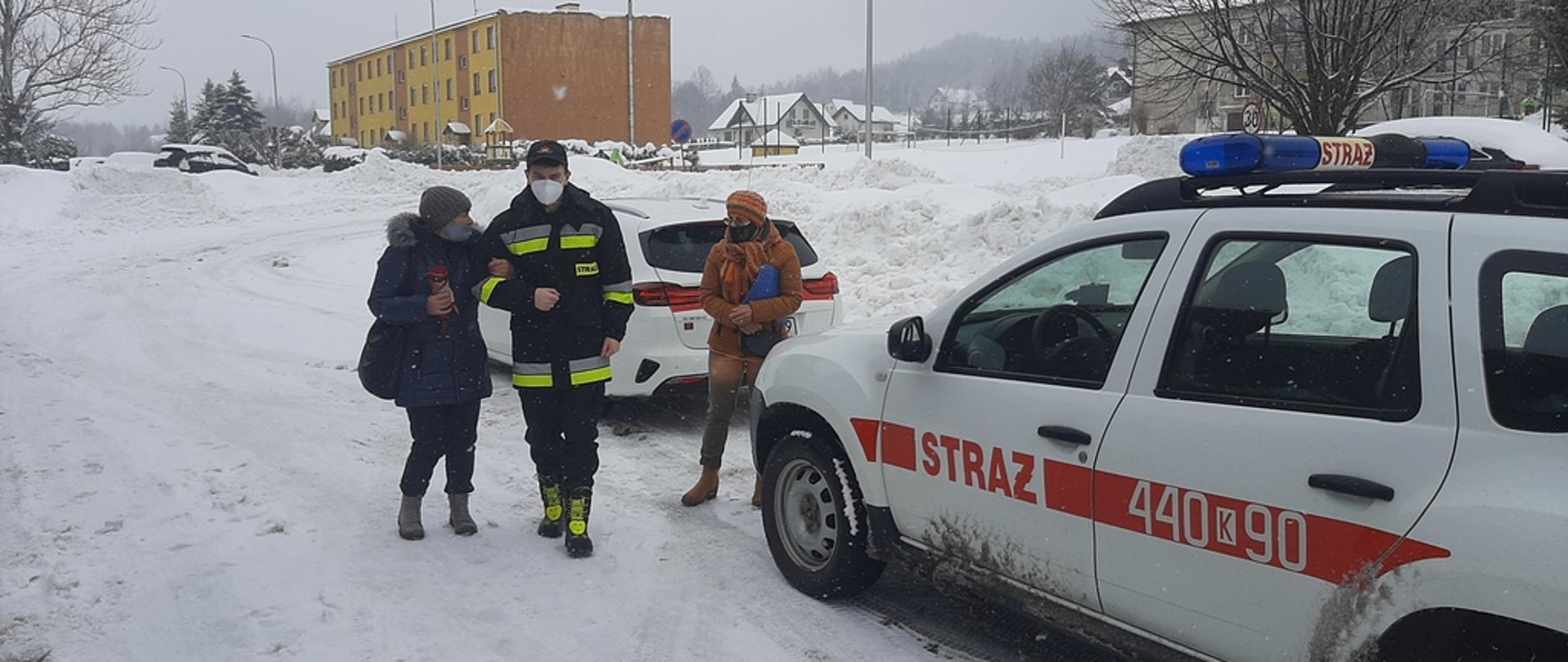 Na zdjęciu widoczny strażak na zaśnieżonej drodze prowadzący kobietę pod rękę do samochodu strażackiego koloru białego którym kobieta zostanie przetransportowana do punktu szczepień. W oddali widoczne bloki mieszkalne.