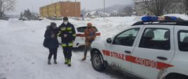 Na zdjęciu widoczny strażak na zaśnieżonej drodze prowadzący kobietę pod rękę do samochodu strażackiego koloru białego którym kobieta zostanie przetransportowana do punktu szczepień. W oddali widoczne bloki mieszkalne.
