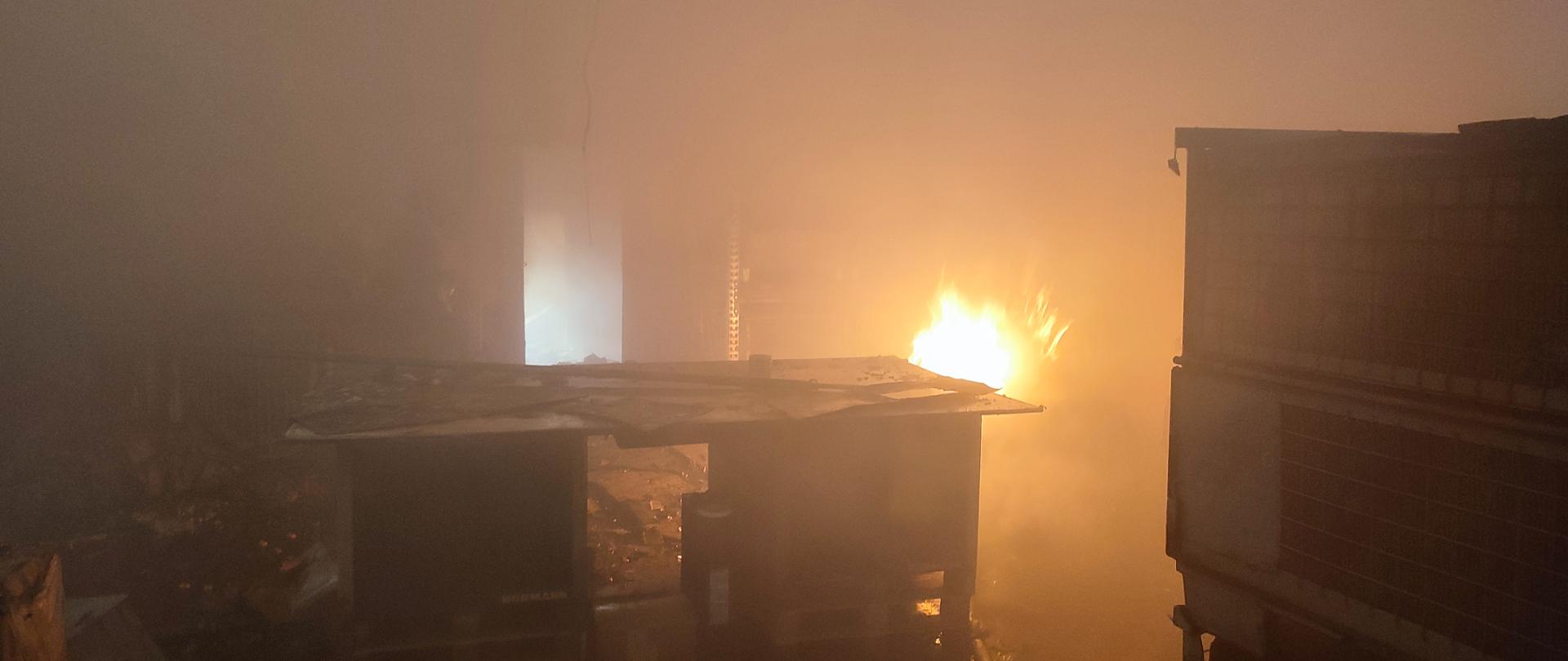 Zdjęcie ukazuje płomień przy regale pomieszczenia, które było objęte pożarem. W pomieszczeniu panuje zadymienie. 
