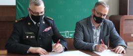 Na zdjęciu dwóch siedzących mężczyzn w maseczkach, jeden w mundurze strażackim drugi w marynarce i koszuli podpisują dokumenty.