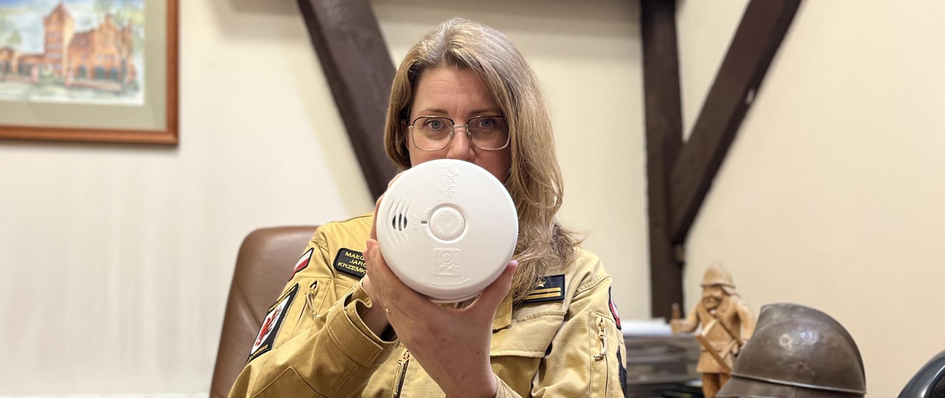 Strażak, kobieta trzyma w ręku czujkę. W tle wyposażenie pokoju.