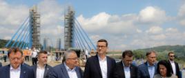 Premier z ministrem infrastruktury i samorządowcami zwiedzają teren budowy mostu.