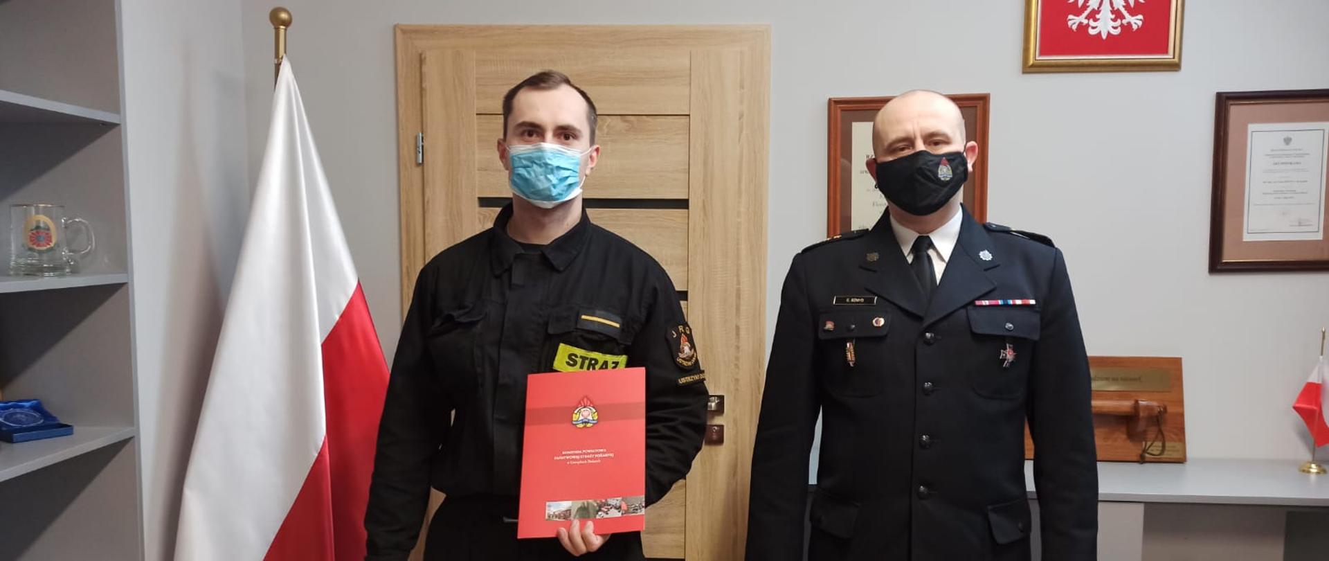 Na zdjęciu widać komendanta powiatowego PSP w Ustrzykach Dolnych w mundurze galowym oraz st. str. Krystiana Nawrockiego który stoi obok w ubraniu koszarowym i w ręce trzyma przekazany od komendanta akt mianowania do służby stałej. Znajdują się Oni w biurze komendanta gdzie w rogu obok drzwi ustawiona jest flaga państwowa a na ścianie wisi godło polski.