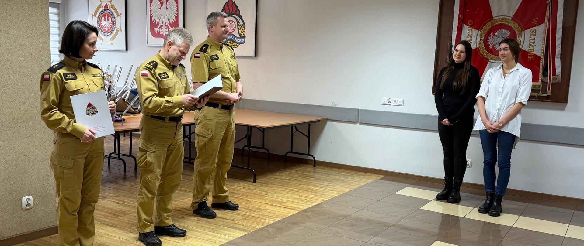 Na zdjęciu widać uroczystą zbiórkę w sali konferencyjnej komendy straży pożarnej. Po lewej stronie stoją funkcjonariusze w mundurach koloru piaskowego – jedna z osób trzyma otwartą teczkę z dokumentem i odczytuje treść decyzji, obok stoi kobieta z okolicznościową teczką z emblematem PSP.
W tle, przy ścianie, stoją dwie kobiety w stroju cywilnym. Na ścianach widoczne są godło Polski, emblemat straży pożarnej oraz sztandar jednostki w gablocie. Wnętrze jest jasne, z kafelkową podłogą i stołami ustawionymi pod ścianą, co podkreśla oficjalny charakter spotkania.