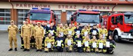 Na zdjęciu widać grupę strażaków stojących przed budynkiem Państwowej Straży Pożarnej. Za nimi ustawione są trzy czerwone wozy strażackie.
Grupa składa się z kilkudziesięciu osób — część ubrana jest w jasnożółte mundury bojowe z odblaskowymi pasami, a część w piaskowe mundury służbowe. Wszyscy stoją lub klęczą w kilku rzędach i trzymają w rękach dyplomy.