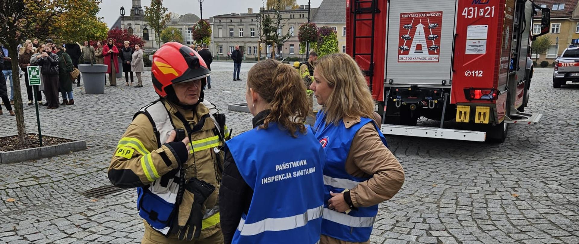 Dowódca akcji przeprowadza rozmowę z przedstawicielami Sanepidu. W tle stoi pojazd pożarniczy.