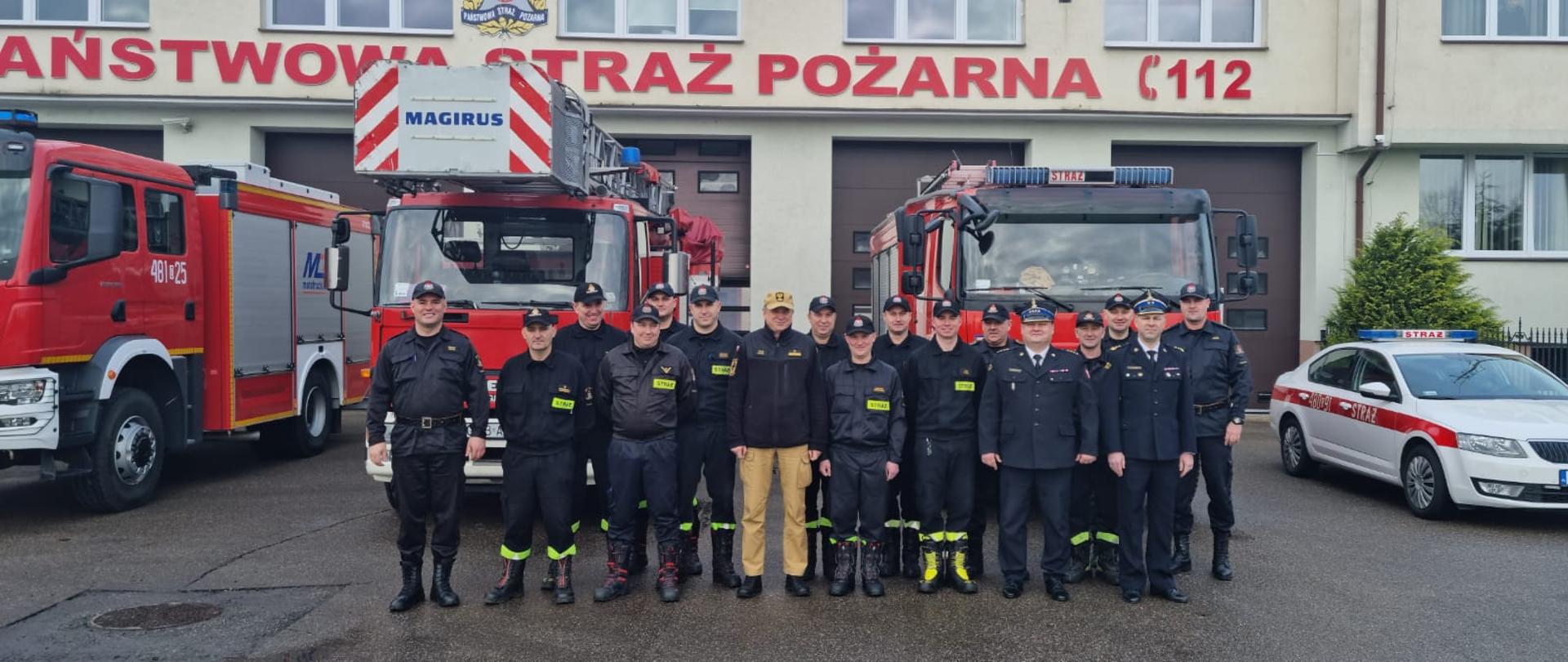 Strażacy stoją obok siebie za nimi przed budynkiem stoją samochody gaśnicze. 