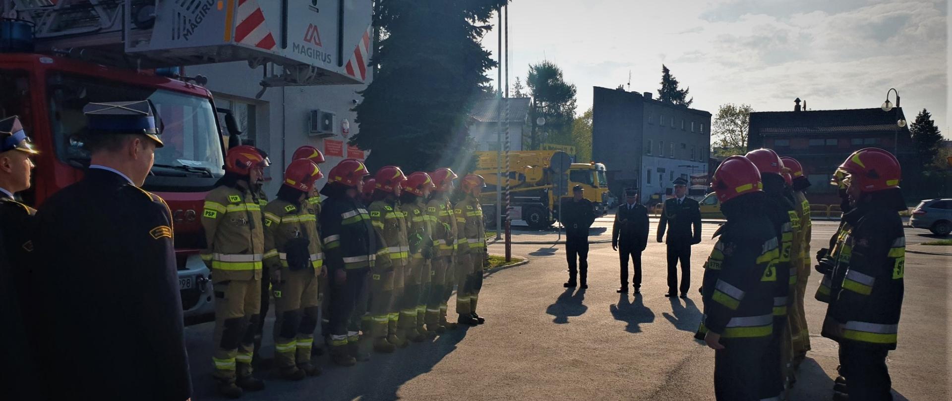 Dwie grupy strażaków w umundurowaniu stojące naprzeciwko siebie. Na środku trzech strażaków w umundurowaniu galowym – Komendant Powiatowy Państwowej Straży Pożarnej w Radomsku oraz jego Zastępca.