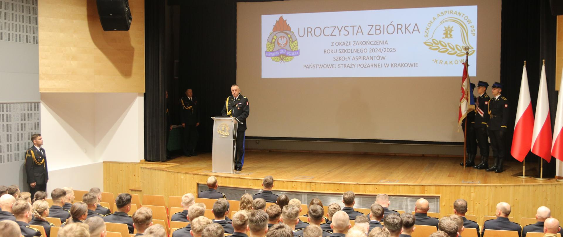 Nadbryg. Piotr Filipek w czasie okolicznościowego przemówienia – mężczyzna stojący za ustawionym na scenie auli pulpitem 