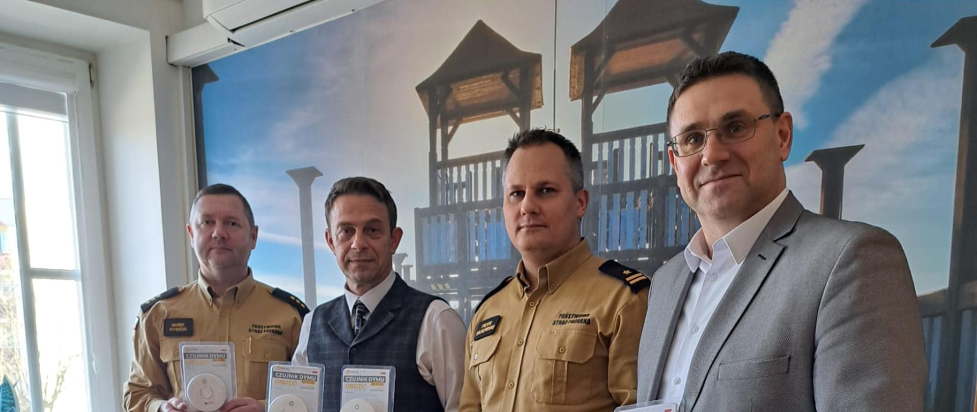 Czterej mężczyźni w postawie stojącej, którzy w dłoniach trzymają i prezentują czujki czadu i dymu.