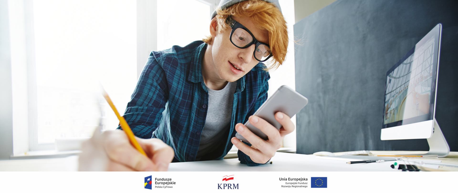 Młody chłopak (nastolatek) oparty o biurko, pisze coś ołówkiem. W tym samym czasie patrzy na ekran smartfona, w tle - włączony komputer.