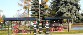 Zdjęcie przedstawia trzech strażaków w strojach bojowych podnoszących flagę państwową na maszt, w tle widać dwa zabytkowe wozy bojowe i cztery drzewa