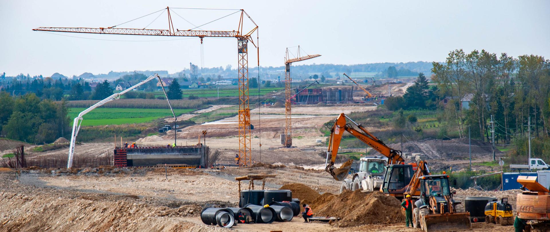 Na zdjęciu przedstawiono plac budowy drogi ekspresowej w otwartym terenie. W centralnej części kadru widoczne są duże maszyny budowlane, w tym koparka wykonująca prace ziemne oraz walec drogowy przygotowujący podłoże. Obok nich znajdują się pracownicy w odzieży roboczej, którzy nadzorują prace.
W tle rozciąga się szeroki pas przygotowywanego nasypu, na którym widać kilka wysokich żurawi wieżowych oraz betonowe konstrukcje – prawdopodobnie elementy przyszłych wiaduktów lub mostów. Na dalszym planie widoczne są zielone pola i drzewa, co podkreśla kontrast między naturalnym krajobrazem a intensywnymi pracami budowlanymi.
Kolorystyka zdjęcia jest naturalna, z dominacją odcieni ziemi (brązy, beże) oraz zieleni w tle. Niebo jest jasne, lekko zachmurzone, co sugeruje neutralne warunki pogodowe sprzyjające pracom.