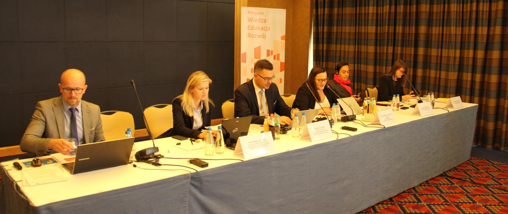 Sześć osób przy długim stole przykrytym materiałem, za nimi roll-up- z napisem Program Wiedza Edukacja Rozwój
