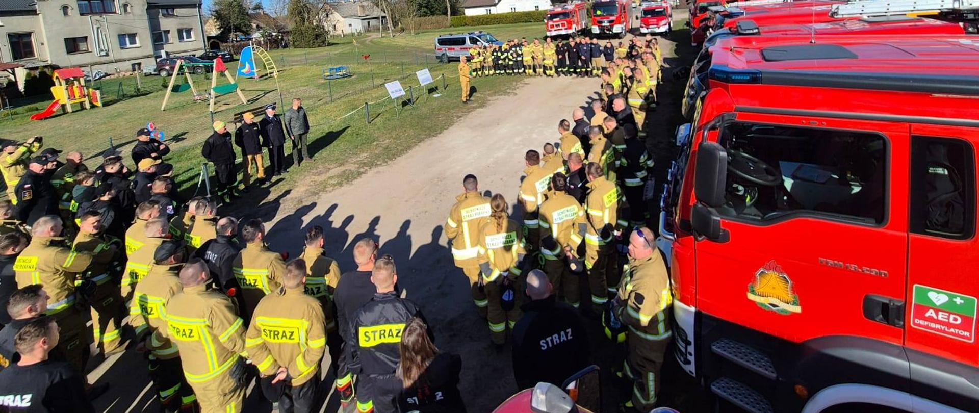 Zbiórka przed warsztatami. Strażacy w dużej ilości stojący przed samochodami.