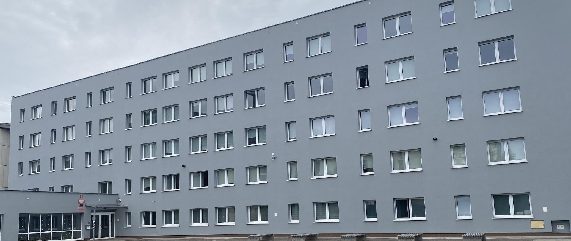 Zdjęcie przedstawia popielaty budynek bursy z licznymi oknami, po lewj stronie widać drzw wejsciowe, przed budynkiem znajdują się ławki