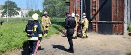 Na zdjęciu widzimy strażaków stawiających drabinę o ścianę ćwiczebną. Strażacy ubrani są w odzież specjalną koloru piaskowego, białe hełmy na głowach.
Jeden ze strażaków trzyma wąż strażacki i szykuje się do wejścia na drabinę.
