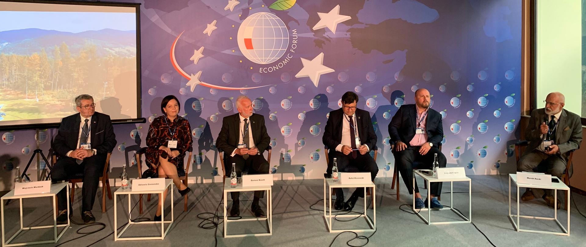 Uczestnicy forum siedzą w rzędzie na krzesłach, za nimi kolorowa ściana.