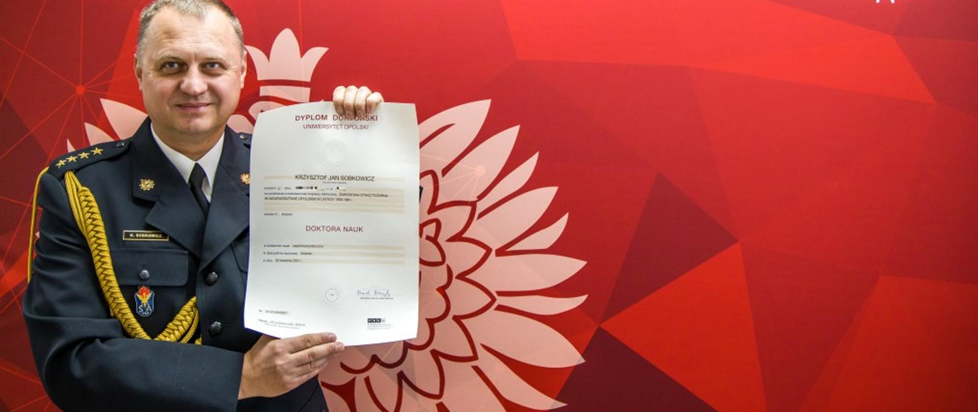 St. kpt. dr Krzysztof Sobkowicz pokazujący dyplom doktorski Uniwersytetu Opolskiego po uzyskaniu stopnia naukowego doktora nauk humanistycznych. Strażak w ubraniu galowym mający w tle planszę koloru czerwonego z białym godłem i napisem Państwowa Straż Pożarna i Komenda Wojewódzka PSP w Opolu.