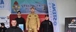 Strażacy stojący na podium po zawodach strażackich. Trzymają dyplomy, puchary oraz nagrody.