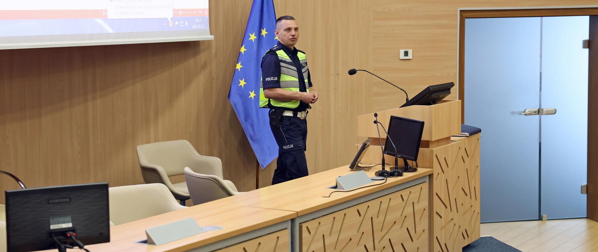Policjant z Wydziału Ruchu Drogowego Komendy Miejskiej Policji w Częstochowie prowadzi szkolenie stojąc przy mównicy w auli CS PSP. W tle widoczna jest flaga Unii Europejskiej oraz ekran multimedialny