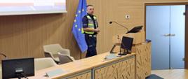 Policjant z Wydziału Ruchu Drogowego Komendy Miejskiej Policji w Częstochowie prowadzi szkolenie stojąc przy mównicy w auli CS PSP. W tle widoczna jest flaga Unii Europejskiej oraz ekran multimedialny