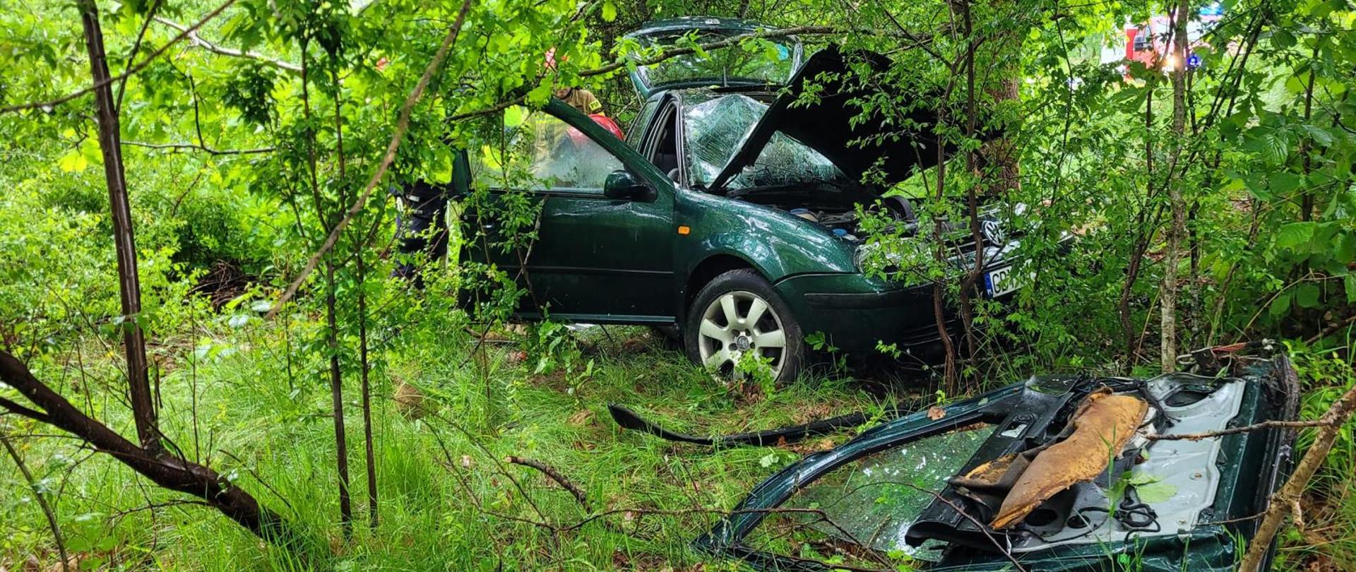 Zielone auto rozbite w lesie, ma otwarte drzwi, maskę oraz bagażnik. Jedne drzwi z auta leżą przed rozbitym samochodem