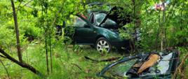 Zielone auto rozbite w lesie, ma otwarte drzwi, maskę oraz bagażnik. Jedne drzwi z auta leżą przed rozbitym samochodem
