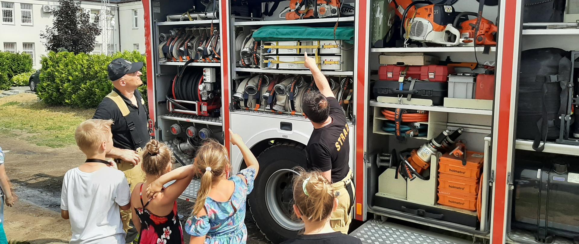 Zdjęcie obrazuje dwóch strażaków ubranych w czarne koszulki typu T-shirt i spodnie specjalne w kolorze piaskowym. Strażacy omawiają sprzęt pożarniczy czterem dzieciom, jednemu chłopcu i trzem dziewczynkom, które patrzą się w stronę samochodu strażackiego. Pojazd ma otwarte boczne skrytki, w których są poukładane różne sprzęty ratownicze i gaśnicze. W otwartych skrytkach widać poukładane w rzędach węże strażackie, pomarańczowo-białe piły spalinowe do cięcia drewna oraz sprzęt hydrauliczny w postaci nożyc i rozpieraka używany podczas uwalniania poszkodowanych w zdarzeniach drogowych.