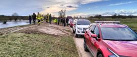 Fotografia przestawia ujęcie wzdłuż wału przeciwpowodziowego, na drodze przy wale zaparkowane pojazdy osobowe (w tym straży pożarnych). Uczestnicy przeglądu wałów idą po koronie wału w kierunku wykonującego fotografię.