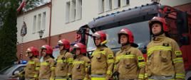 Siedmiu strażaków w umundurowaniu specjalnym oraz hełmach stoi na baczność w szeregu przed samochodami ratowniczo-gaśniczymi. Samochody ustawione są przed garażami od strony ulicy Armii Krajowej, mają włączone sygnały swietlne i dzwiękowe. Dowódca salutuje