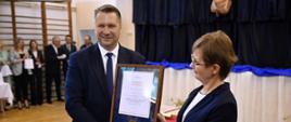 Minister Czarnek i kobieta w granatowej marynarce i okularach stoją obok siebie, razem trzymają oprawiony w ramki dokument, za nimi przysłonięta biało-granatową kotarą ściana, z boku w tle pod ścianą z drabinkami stoi szereg osób.