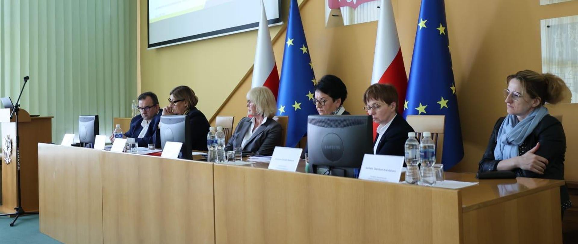 Narada dotycząca funkcjonowania systemu Państwowe Ratownictwo Medyczne na terenie województwa opolskiego. Na zdjęciu od lewej siedzą: Dyrektor Opolskiego Centrum Ratownictwa Medycznego w Opolu Jarosław Kostyła, Dyrektor Opolskiego Oddziału NFZ Beata Cyganiuk, Dyrektor Wydziału Zdrowia OUW Mirosława Kwatek-Hoffmann, Wojewoda Opolski Monika Jurek, Wicemarszałek Województwa Opolskiego Zuzanna Donath-Kasiura oraz Dyrektor Departamentu Zdrowia i Polityki Społecznej w Urzędzie Marszałkowskim Województwa Opolskiego Izabela Damboń-Kandziora. Za nimi widać flagi Polski oraz Unii Europejskiej. W lewym, górnym rogu widać omawianą prezentację. 