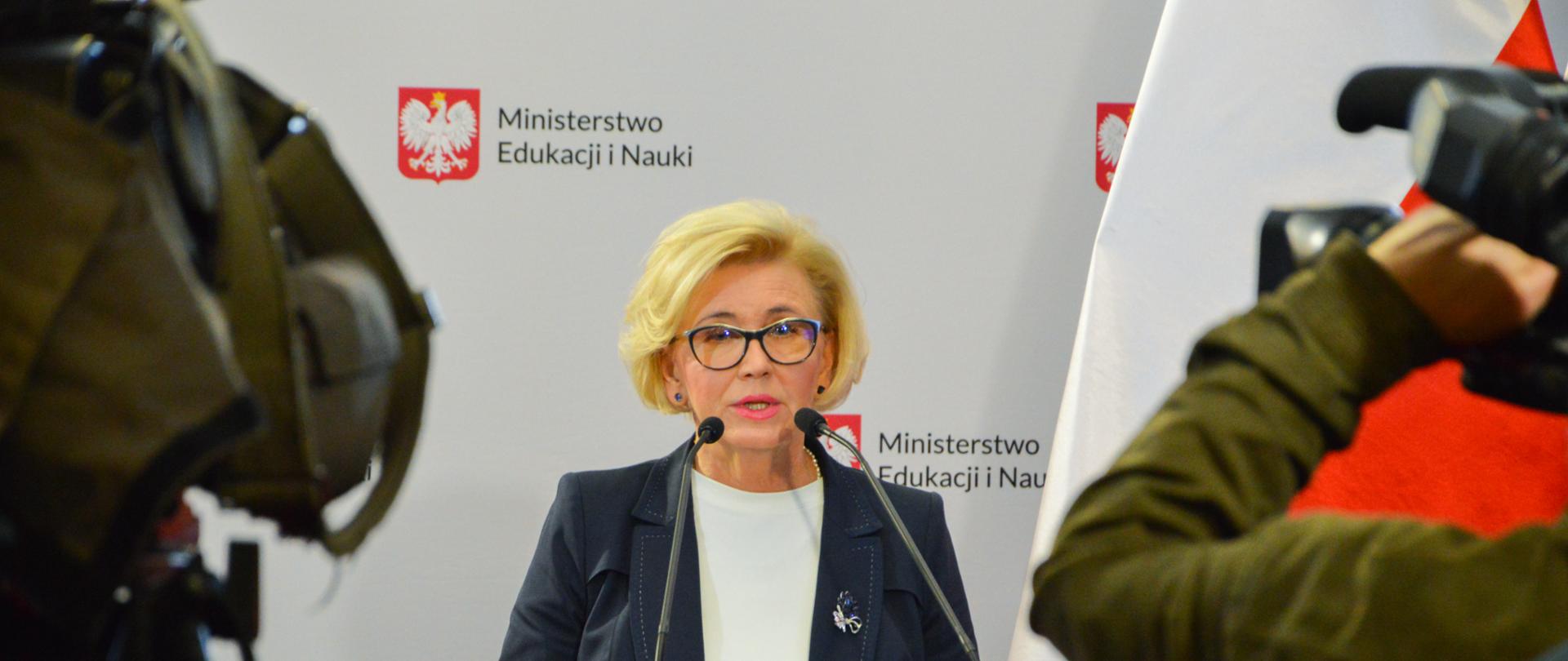 Wiceminister stoi przy mównicy z orłem, w tle biało-czerwona flaga. Na pierwszym planie kamery 