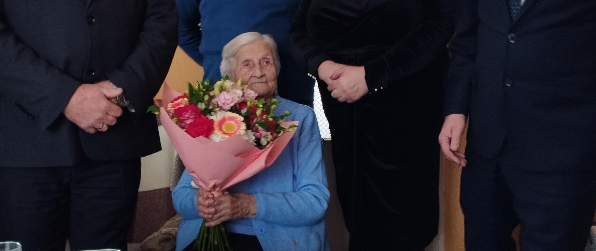 Na krześle, pośrodku pomieszczenia, siedzi starsza kobieta w niebieskim swetrze. Za jej plecami stoją cztery elegancko ubrane osoby: trzech mężczyzn i kobieta. Jubilatka trzyma w rękach bukiet kolorowych kwiatów. Jest przejęta podniosłą chwilą i delikatnie się uśmiecha. Przed Jubilatką stoi świątecznie nakryty stół z tortem i słodkościami