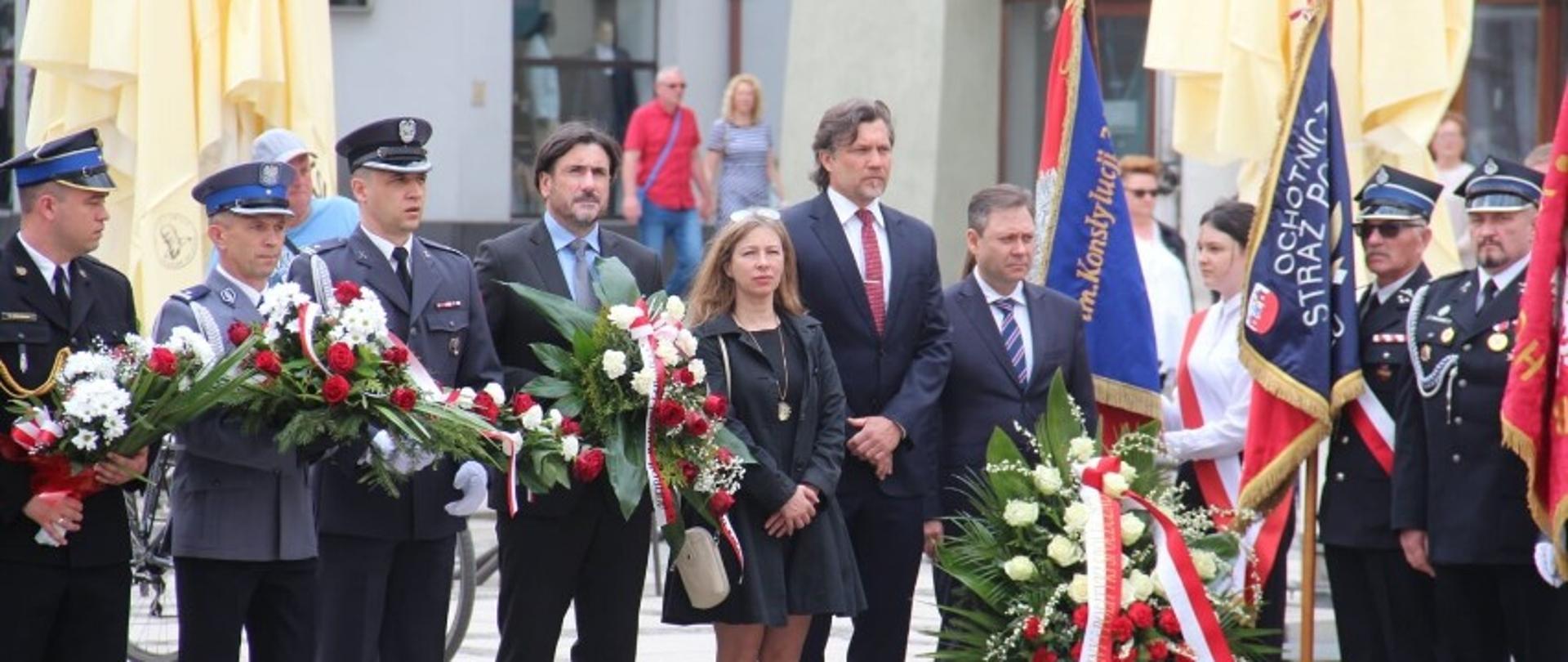 Na zdjęciu widać funkcjonariuszy w umundurowaniu wyjściowym. Wśród funkcjonariuszy strażak, policjant i inne służby. Zdjęcie zrobione w ciągu dnia, w czasie uroczystości. Uroczystość ma miejsce przed kaliskim ratuszem. Na zdjęciu widać poczty sztandarowe.