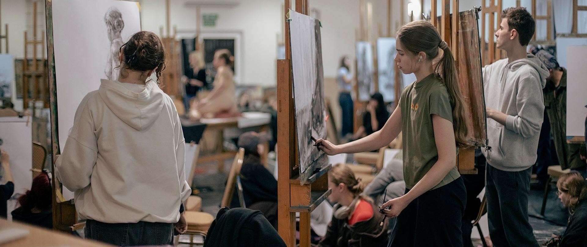 Na zdjęciu widać dużą, oświetloną pracownię artystyczną. Na pierwszym planie, z lewej strony, widać osobę w białej bluzie z kapturem, która stoi przed sztalugami i rysuje na dużym płótnie. Z prawej strony, bardziej na środku, inna osoba w zielonej koszulce również rysuje, a obok niej stoi młody mężczyzna w szarej bluzie. W tle widocznych jest wiele innych osób w różnym wieku, które rysują lub przyglądają się pracom, co nadaje zdjęciu atmosferę wspólnego tworzenia.