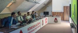 Czterech mężczyzn (pierwszy i ostatni w ubraniu cywilnym, dwaj środkowi w umundurowaniu PSP) - przedstawiciele Komendy Głównej PSP siedzących za stołem konferencyjnym. Na podstawie stołu umieszczony jest baner z napisem i logo Komendy Głównej Państwowej Straży Pożarnej. 