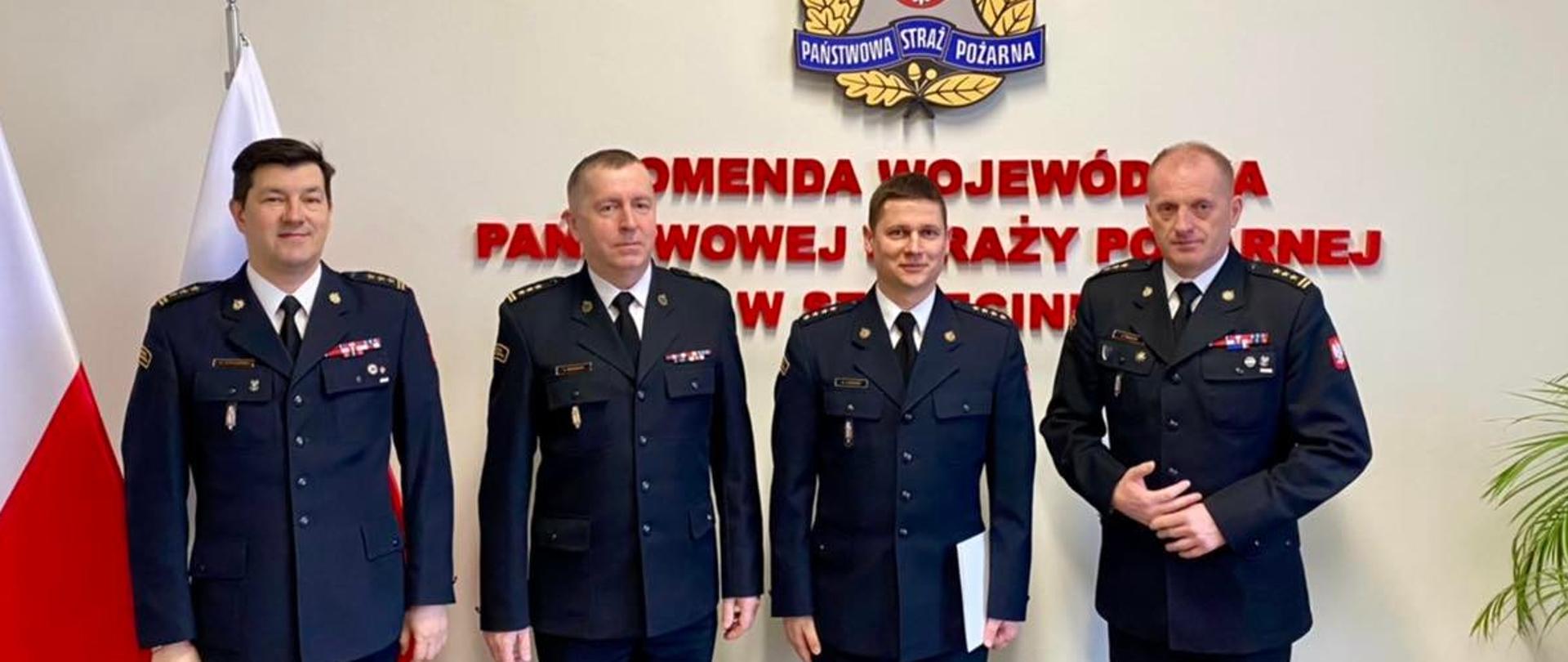 Czterech strażaków na tle napisu Komenda Wojewódzka PSP w Szczecinie, w tle logo PSP oraz dwie flagi państwowe.
