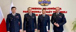 Czterech strażaków na tle napisu Komenda Wojewódzka PSP w Szczecinie, w tle logo PSP oraz dwie flagi państwowe.
