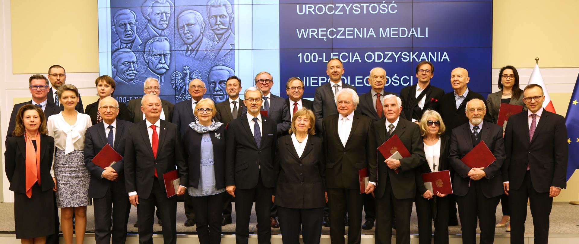 zdjęcie grupowe uczestnicy uroczystości stoją a za nimi napis uroczystość wręczenia medali