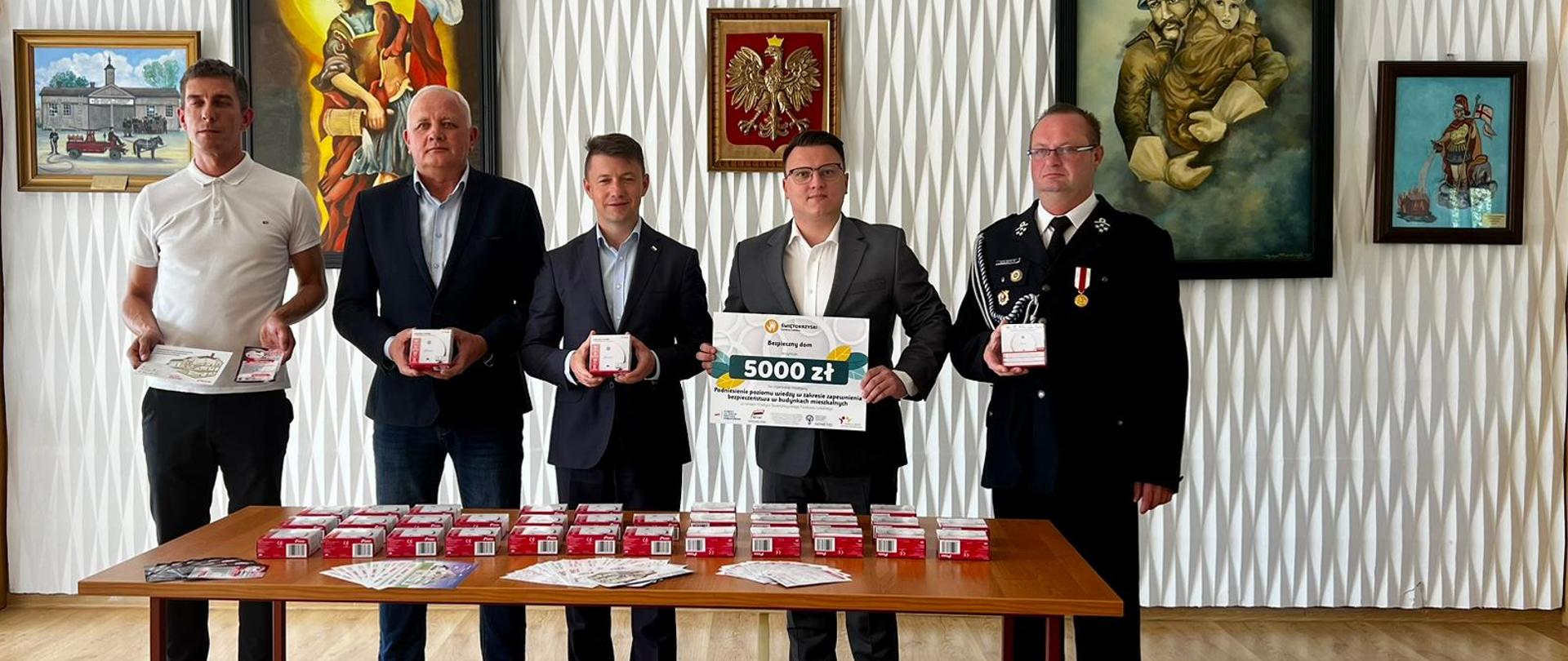 Na stoliku leżą detektory dymu oraz materiały promocyjne akcji, na drugim planie uczestnicy konferencji prasowej 