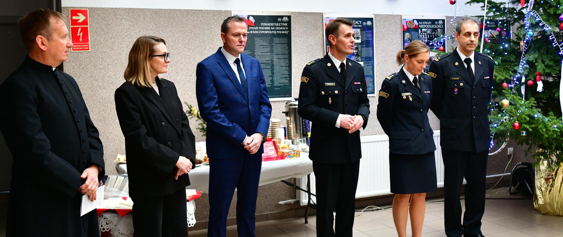 Grupa sześciu osób stoi w jasnym korytarzu obok udekorowanej choinki; z lewej strony mężczyzna w czarnej sutannie, obok niego kobieta w ciemnym garniturze, dalej mężczyzna w niebieskim garniturze, a po prawej trzech funkcjonariuszy w granatowych mundurach strażackich, na ścianie za nimi wiszą tablice informacyjne.