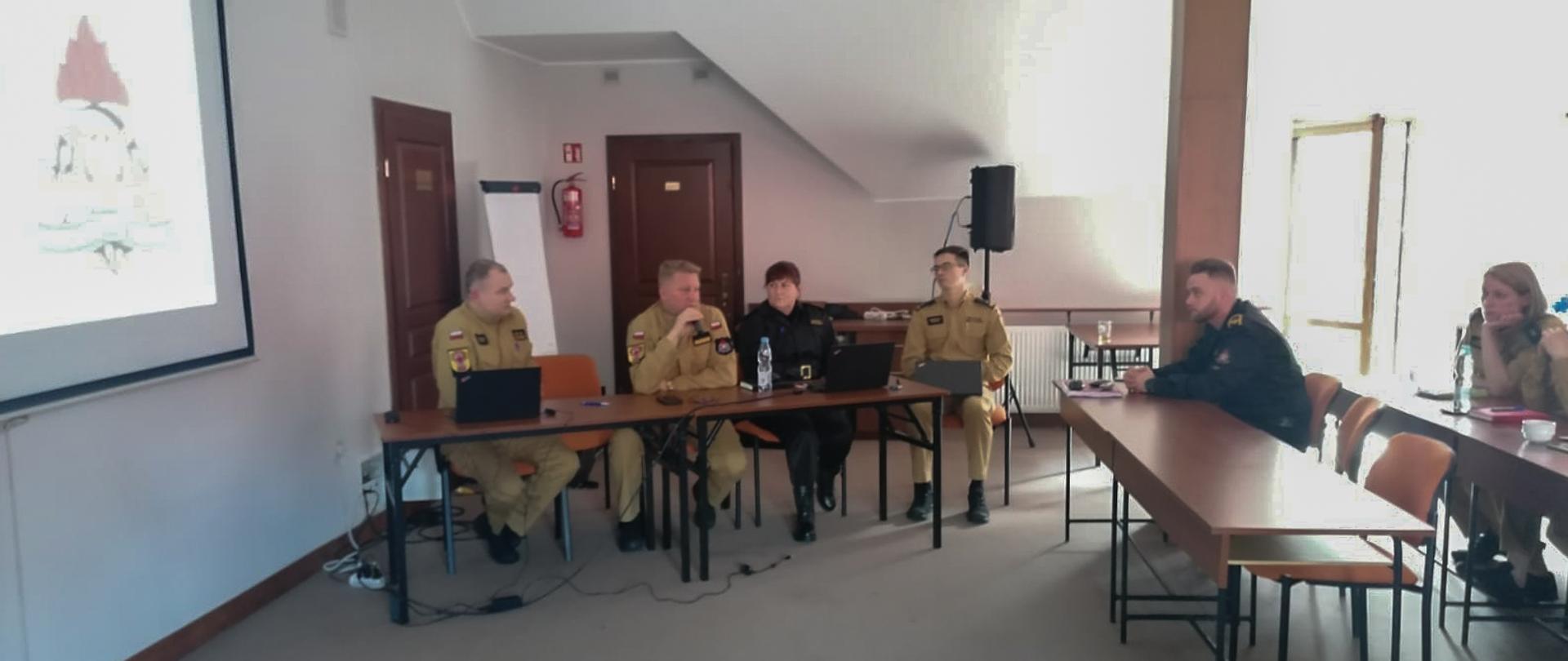 sześcioro strażaków w trakcie szkolenia w sali, 4 siedzi przy poprzecznym stole 2 przy innych