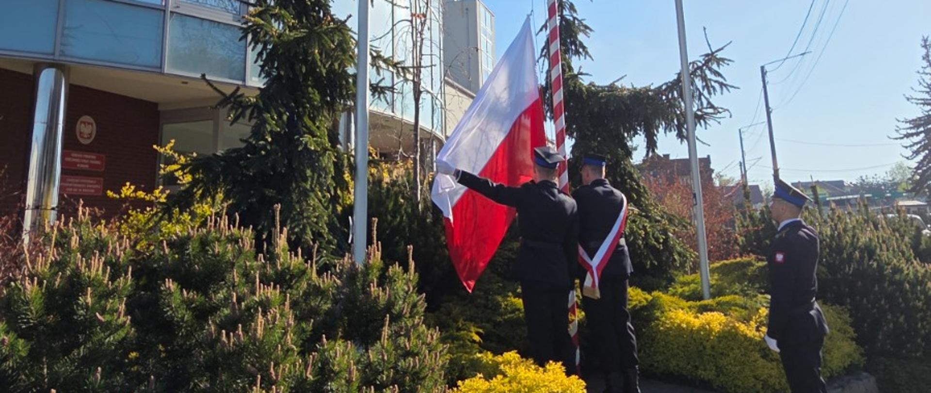 Na zdjęciu widać trzech strażaków stojących przy maszcie flagowym przed budynkiem straży pożarnej. W bliskim otoczeniu masztu znajdują się krzewy i rośliny.
Dwóch strażaków stoi blisko masztu i podnosi biało-czerwoną flagę Polski. Trzeci strażak stoi obok, nieco z tyłu, zwrócony jest w stronę masztu. Wszyscy mają na sobie ciemne, galowe mundury i czapki. Jeden z nich dodatkowo ma przez lewe ramię przełożony biało-czerwony pas.
Maszt, na który wciągana jest flaga, ma biało-czerwone pasy. Obok stoją jeszcze dwa puste maszty.
Maszt znajduje się na podwyższonym rabacie z kamiennym obrzeżem.
