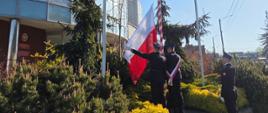 Na zdjęciu widać trzech strażaków stojących przy maszcie flagowym przed budynkiem straży pożarnej. W bliskim otoczeniu masztu znajdują się krzewy i rośliny.
Dwóch strażaków stoi blisko masztu i podnosi biało-czerwoną flagę Polski. Trzeci strażak stoi obok, nieco z tyłu, zwrócony jest w stronę masztu. Wszyscy mają na sobie ciemne, galowe mundury i czapki. Jeden z nich dodatkowo ma przez lewe ramię przełożony biało-czerwony pas.
Maszt, na który wciągana jest flaga, ma biało-czerwone pasy. Obok stoją jeszcze dwa puste maszty.
Maszt znajduje się na podwyższonym rabacie z kamiennym obrzeżem.
