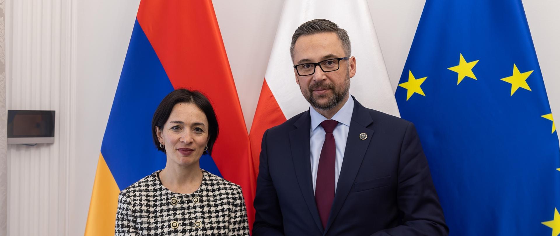 Pod białą ścianą stoi minister Kulasek i minister Zhanną Andreasyan, za nimi pod ścianą flagi Polski, Armenii i UE.