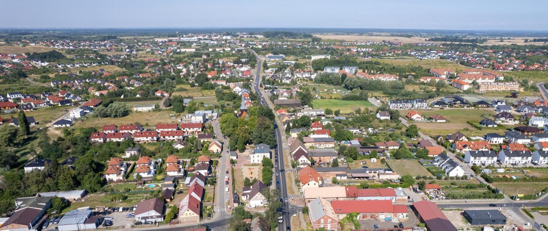 Droga przebiegająca przez miejscowość, po obu stronach liczne zabudowania, pomiędzy nimi drzewa, niebo zachmurzone