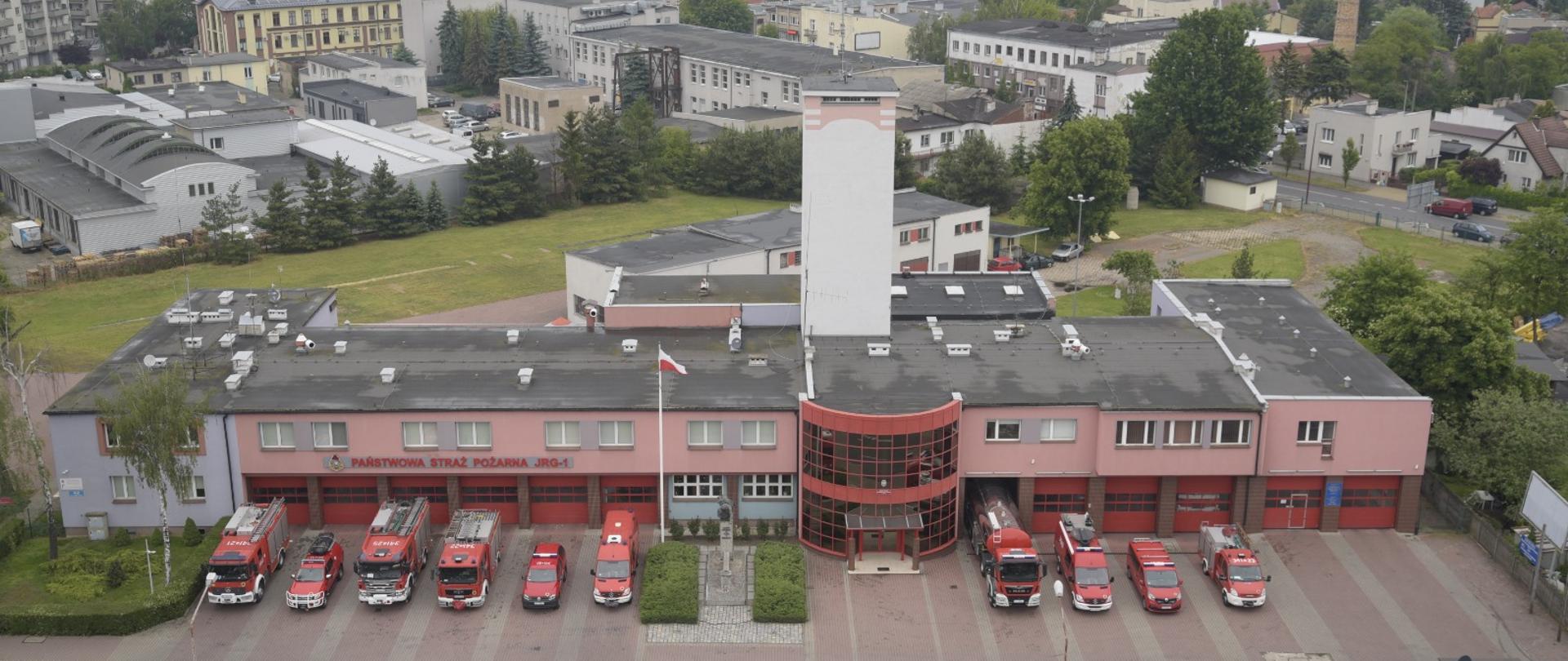 Zdjęcie przedstawia front budynku Komendy Państwowej Straży Pożarnej w Kaliszu oraz Jednostki Ratowniczo Gaśniczej nr 1 w Kaliszu z lotu ptaka. Budynek dwukondygnacyjny koloru łososiowego z wieżą po środku koloru białego. Parter budynku to dwanaście bram wyjazdowych przed którymi wystawione zostały samochody pożarnicze. Pierwsze piętro to linia okien pomieszczeń użytkowych. Na środku budynku widzimy przeszklone wejście główne półokrągłe koloru czerwonego. W dalszym planie widzimy cześć miasta Kalisza - budynki mieszkalne i przemysłowe.
