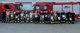 Grupa strażaków pozujących do zdjęcia a w tle wozy strażackie